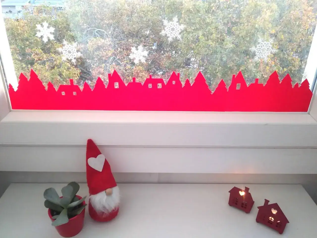 Disegni finestre natale: una silhouette rossa di casette e alberi decora il davanzale, con fiocchi di neve applicati sul vetro.