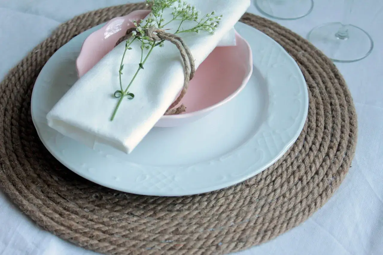 Sottopiatti in stoffa fai da te, con tovagliolo bianco legato da spago e decorato con un rametto fiorito.