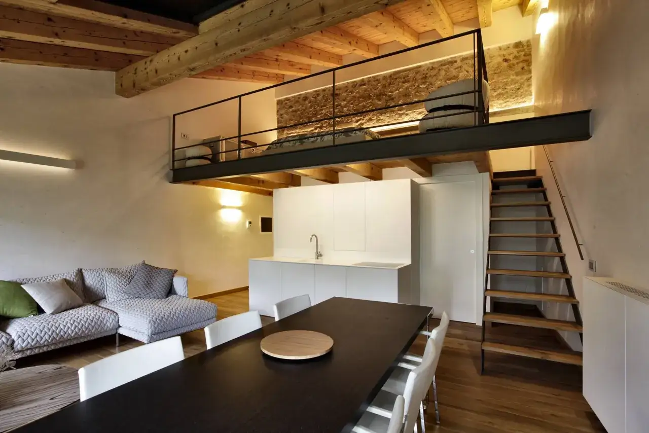 Modern living space with a loft bedroom accessible via stairs. The soppalco soffitto basso features a metal railing and a stone wall.