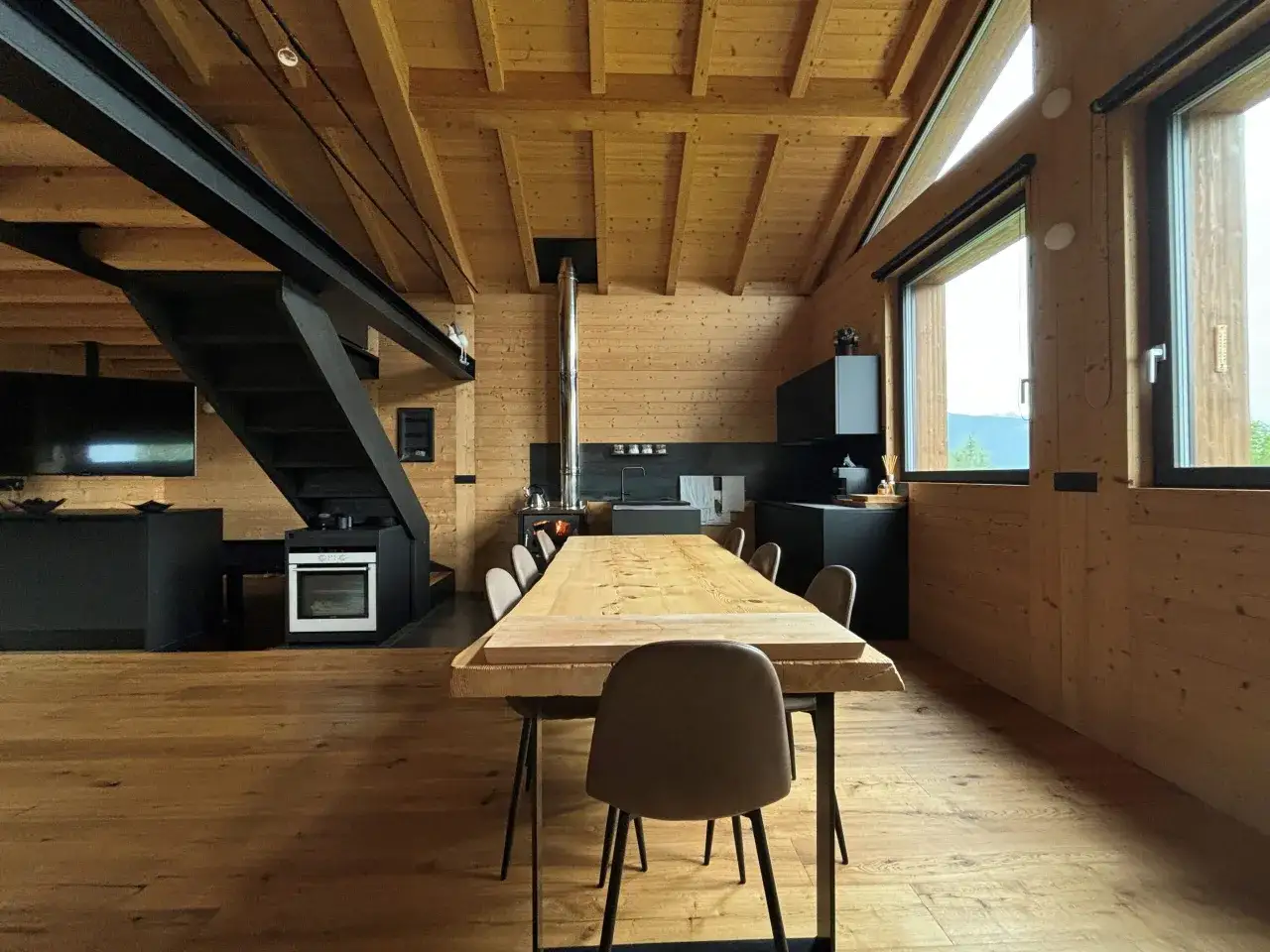 Interno di una casa con tetto in legno a vista, tavolo in legno grezzo, cucina moderna e scala in metallo.