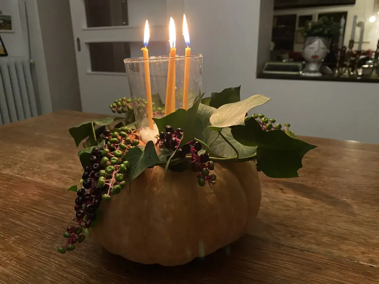 Un semplice centrotavola autunnale con zucca, foglie verdi e bacche scure, illuminato da tre candele gialle che danzano in un bicchiere.