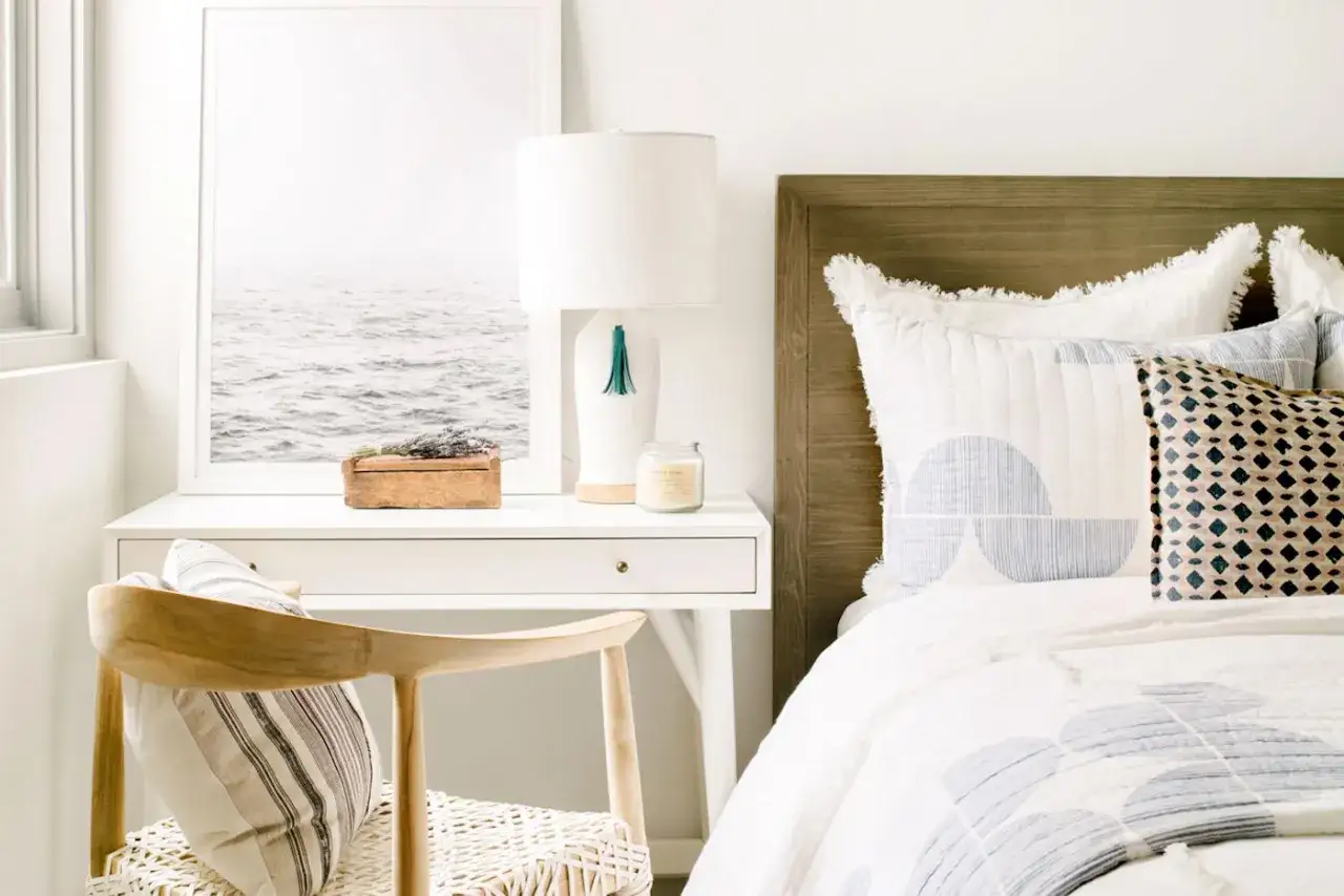 Idee su come disporre i mobili in camera da letto: un comodino bianco con lampada, quadro marino, sedia in legno e letto accogliente.