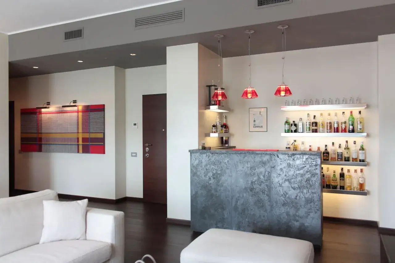 Un angolo bar casa moderno con bancone grigio, bottiglie illuminate su mensole e lampade rosse. Perfetto per creare idee angolo bar casa.