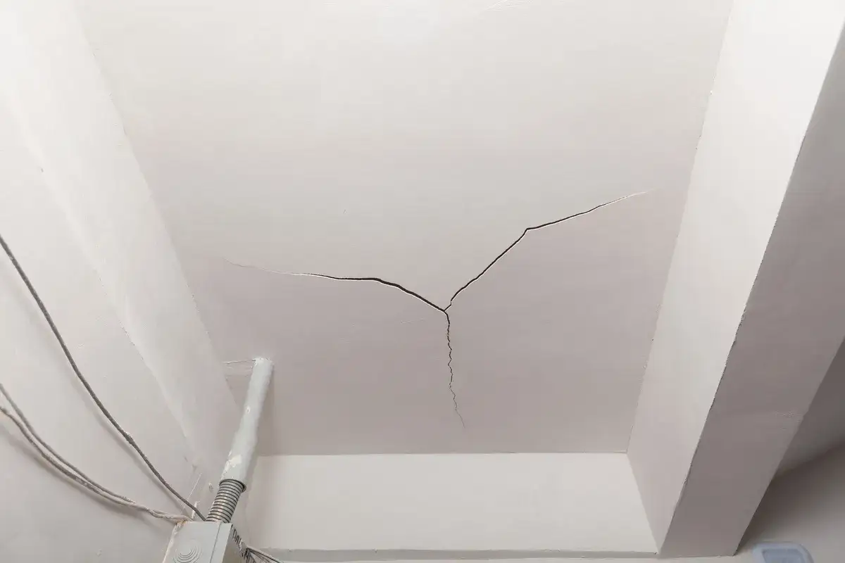 Soffitto bianco con evidenti crepe che si diramano, vicino a tubi e cavi elettrici.