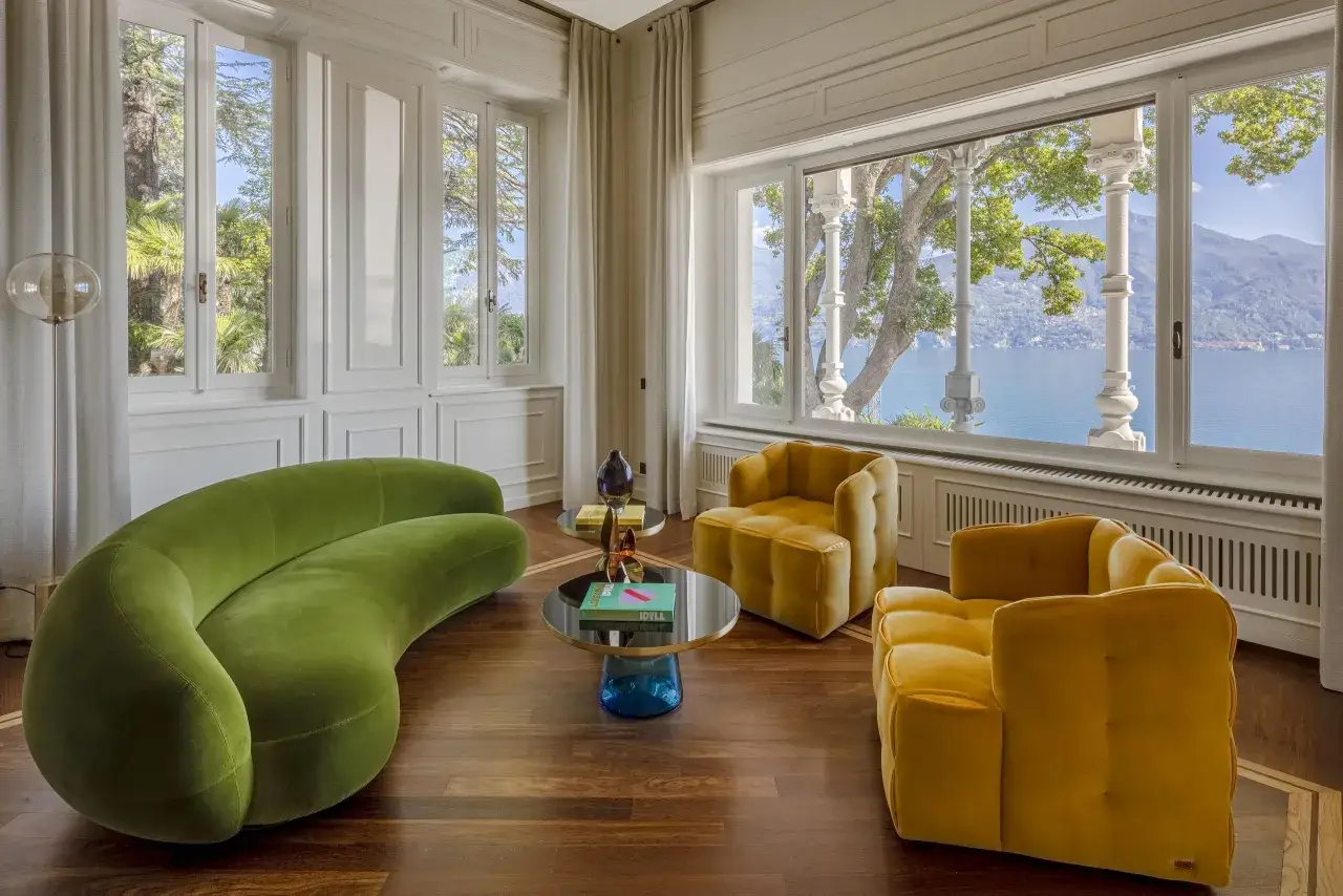 Un salotto con divano verde sinuoso e poltrone gialle, tavolini rotondi e una vista mozzafiato sul lago. L'arredamento in stile art déco crea un'atmosfera elegante e moderna.