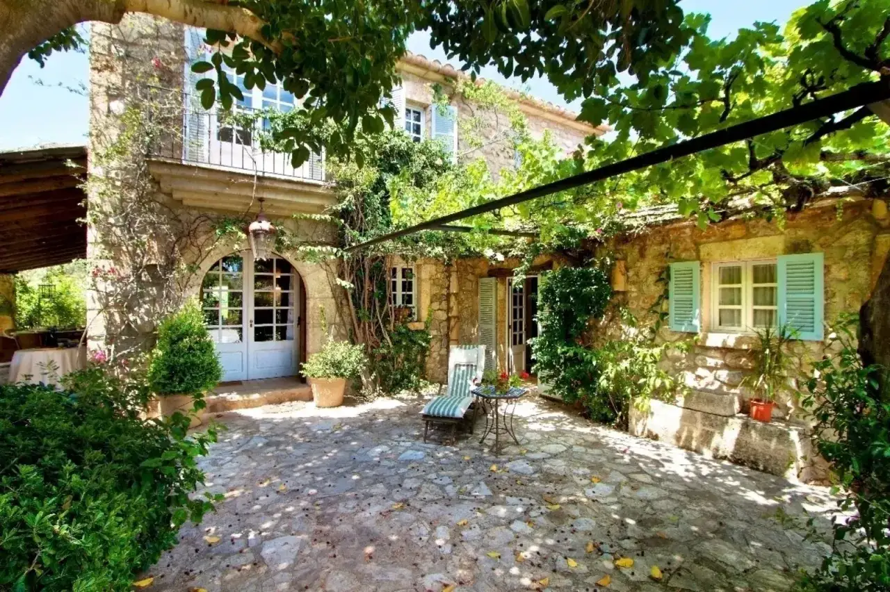 Cortile in pietra con sedie e tavolo, circondato da una casa di campagna in pietra e vegetazione rigogliosa. Idee case di campagna esterni.