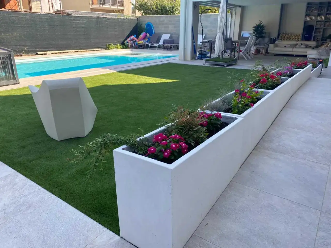 Un cortile in cemento trasformato con fioriere bianche piene di fiori rosa vivaci, prato artificiale e una piscina azzurra.