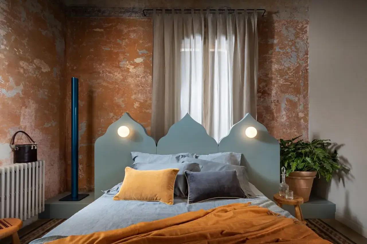 Un letto con testiera a punta, cuscini color ocra e antracite, e una coperta arancione creano un'atmosfera da camere aesthetic. Pareti con texture grezza e dettagli moderni.