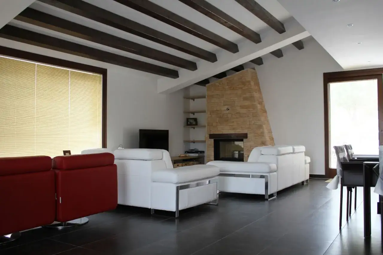 Soggiorno moderno con camino in pietra, divani bianchi e rossi, e un soffitto in cartongesso con travi a vista in legno scuro.
