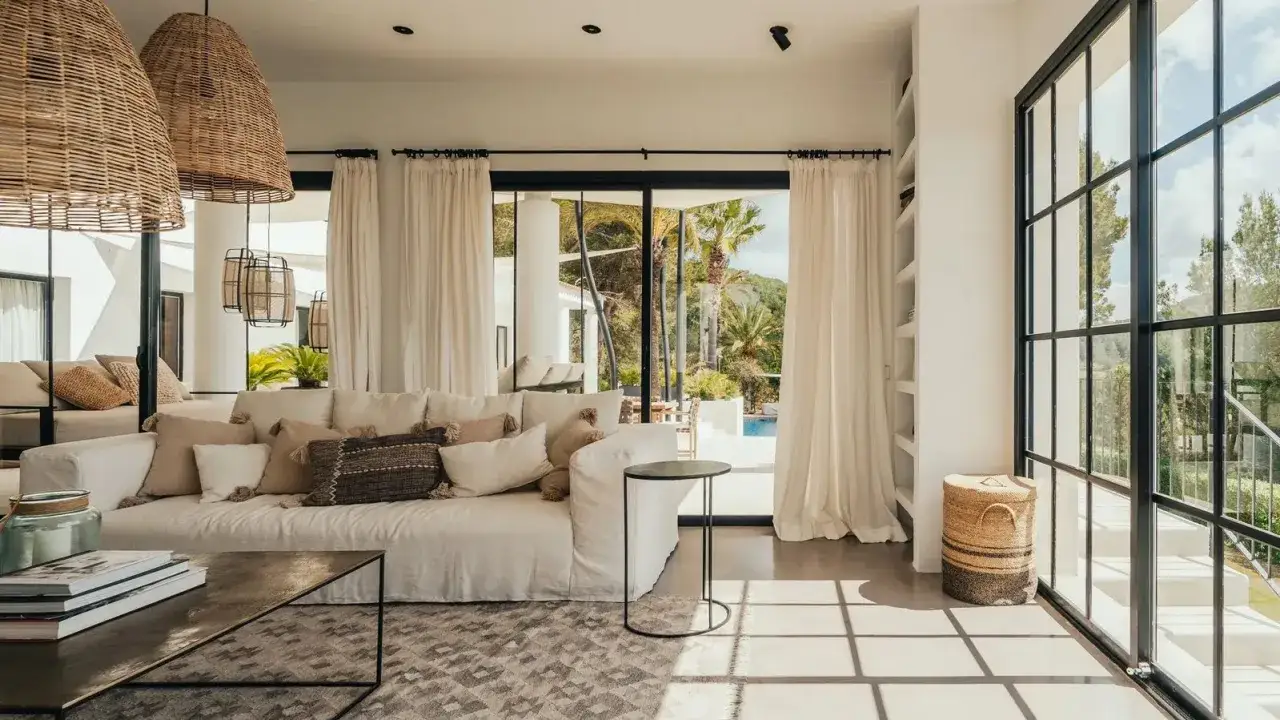 Accogliente salotto casa al mare con divano bianco, cuscini decorativi e grandi vetrate che si affacciano su un giardino lussureggiante.