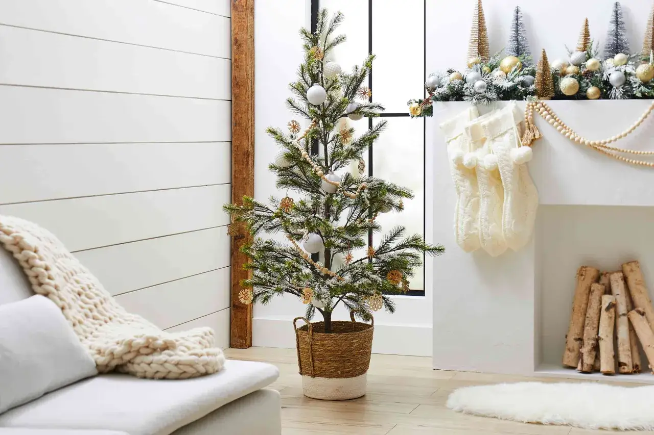Un albero di natale stile scandinavo, decorato con palline bianche e fiocchi di paglia, in un cesto di vimini. Accanto, calze di lana e legna da ardere.