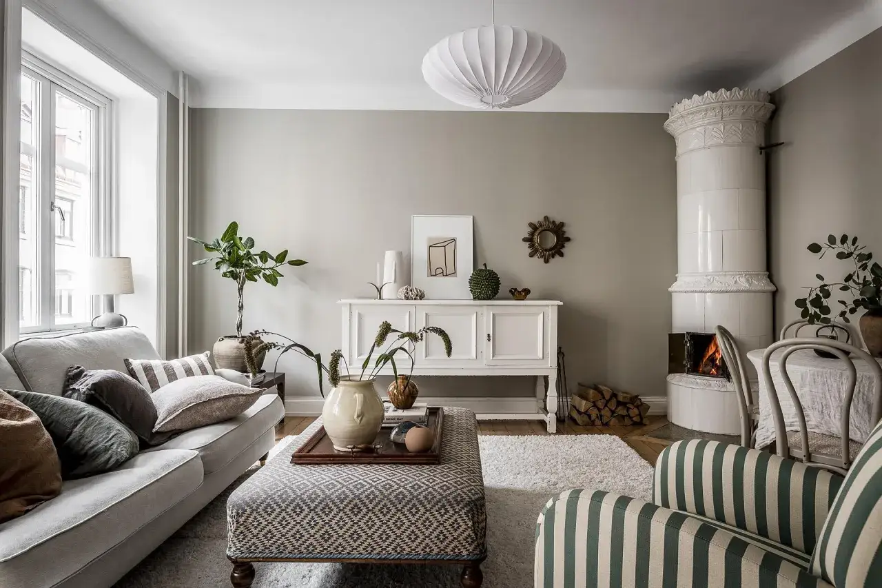 Salotto accogliente con camino in maiolica, divano grigio e pouf decorato. L'arredamento stile classico è completato da un mobile bianco e piante verdi.