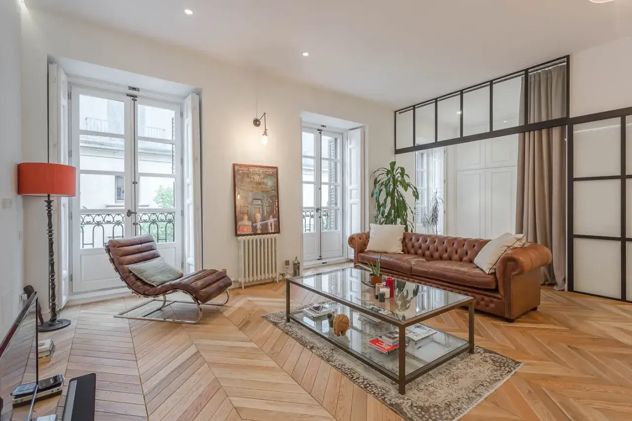 Un soggiorno caldo e accogliente con divano in pelle, chaise longue, tavolino in vetro e parquet a spina di pesce.