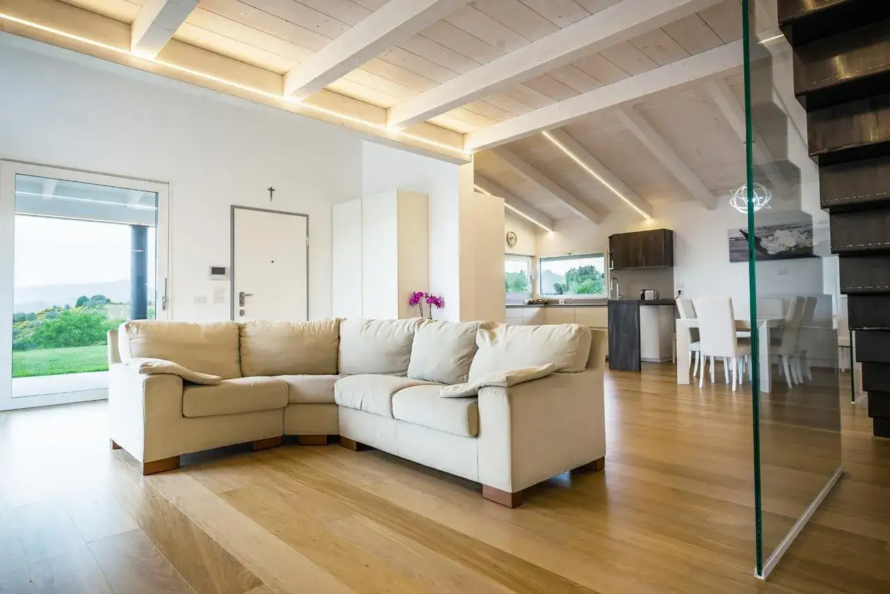 Soggiorno moderno con travi in legno sbiancate senza carteggiare, divano beige, cucina a vista e tavolo da pranzo.
