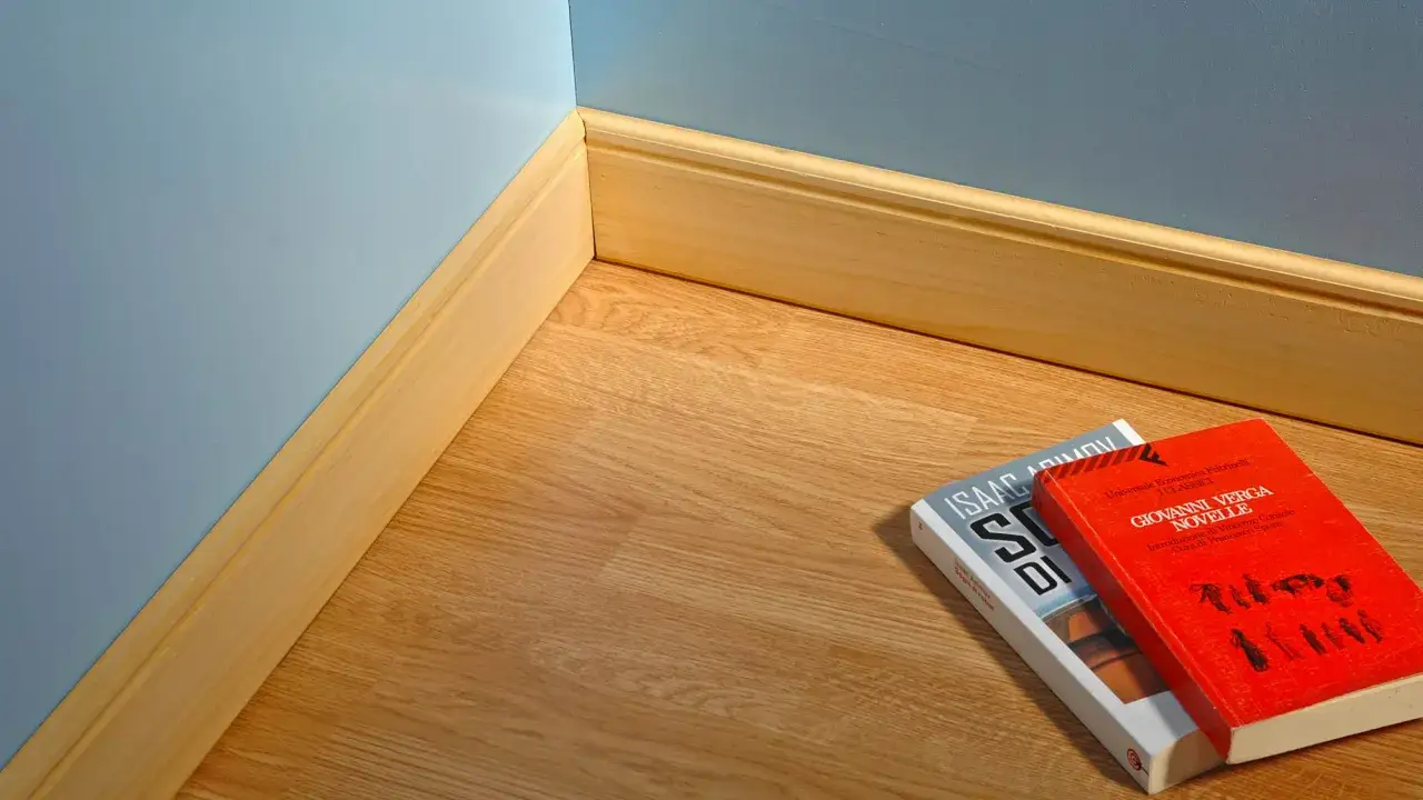 Angolo di una stanza con parquet e pareti azzurre. La posa in opera battiscopa in legno &egrave; curata. Due libri giacciono sul pavimento.
