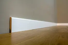Un battiscopa sagomato bianco, con un sottile bordo in legno a vista, corre lungo una parete grigia e un pavimento in parquet lucido.