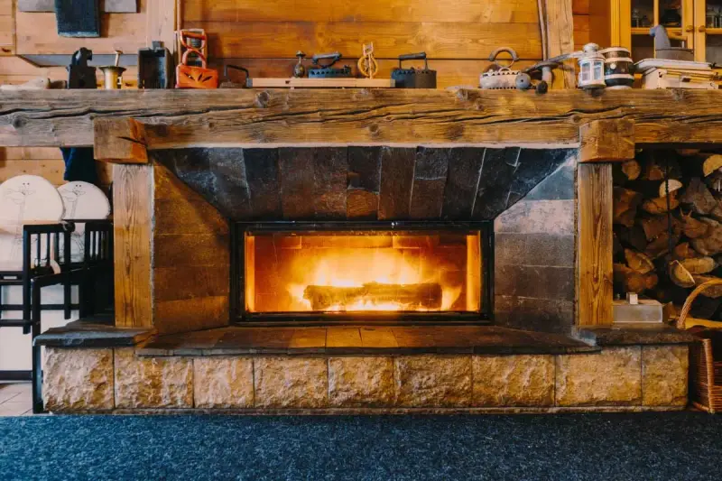 Camino in pietra e legno, con fuoco acceso, che evoca idee camini rustici di montagna. Sopra, oggetti d'epoca e legna accatastata.